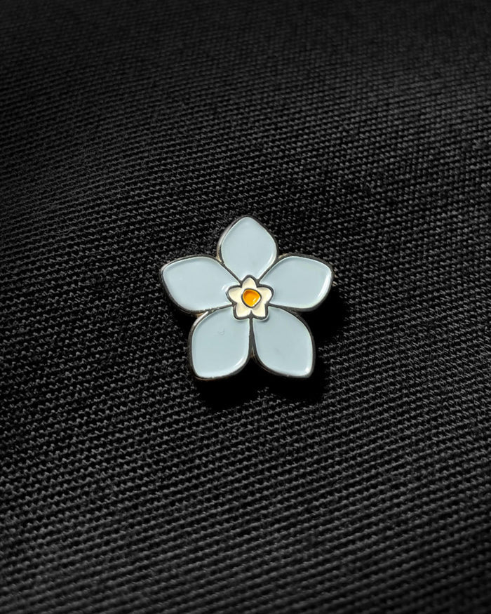Ela Mo™ Mini Pin für Rucksäcke | Vergissmeinnicht. Nahansicht eines kleinen Pins, befestigt auf dem schwarzen Rucksack.