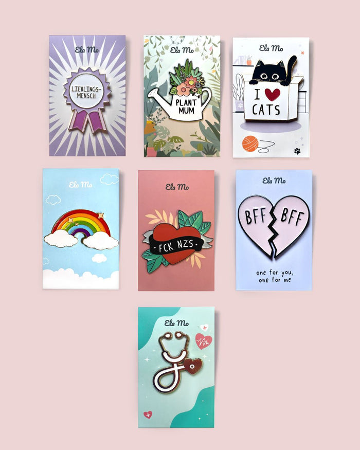 Ela Mo™ Pin für Rucksäcke | Cat. Verschiedene Ausführungen von Rucksackpins auf rosa Hintergrund.